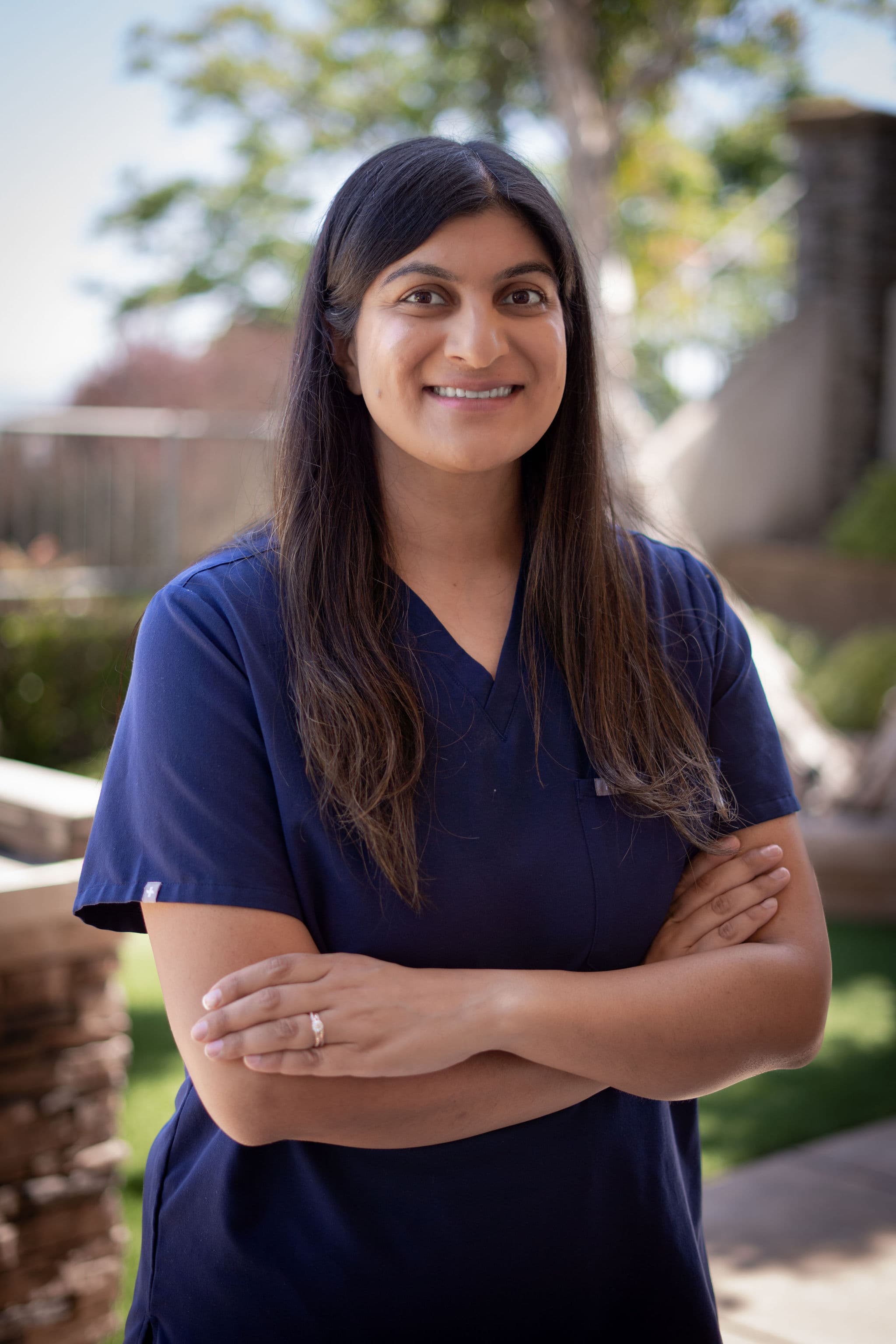 Nicki Rao outdoors in navy scrubs, arms folded, smiling toward the camera with trees and a garden behind her.