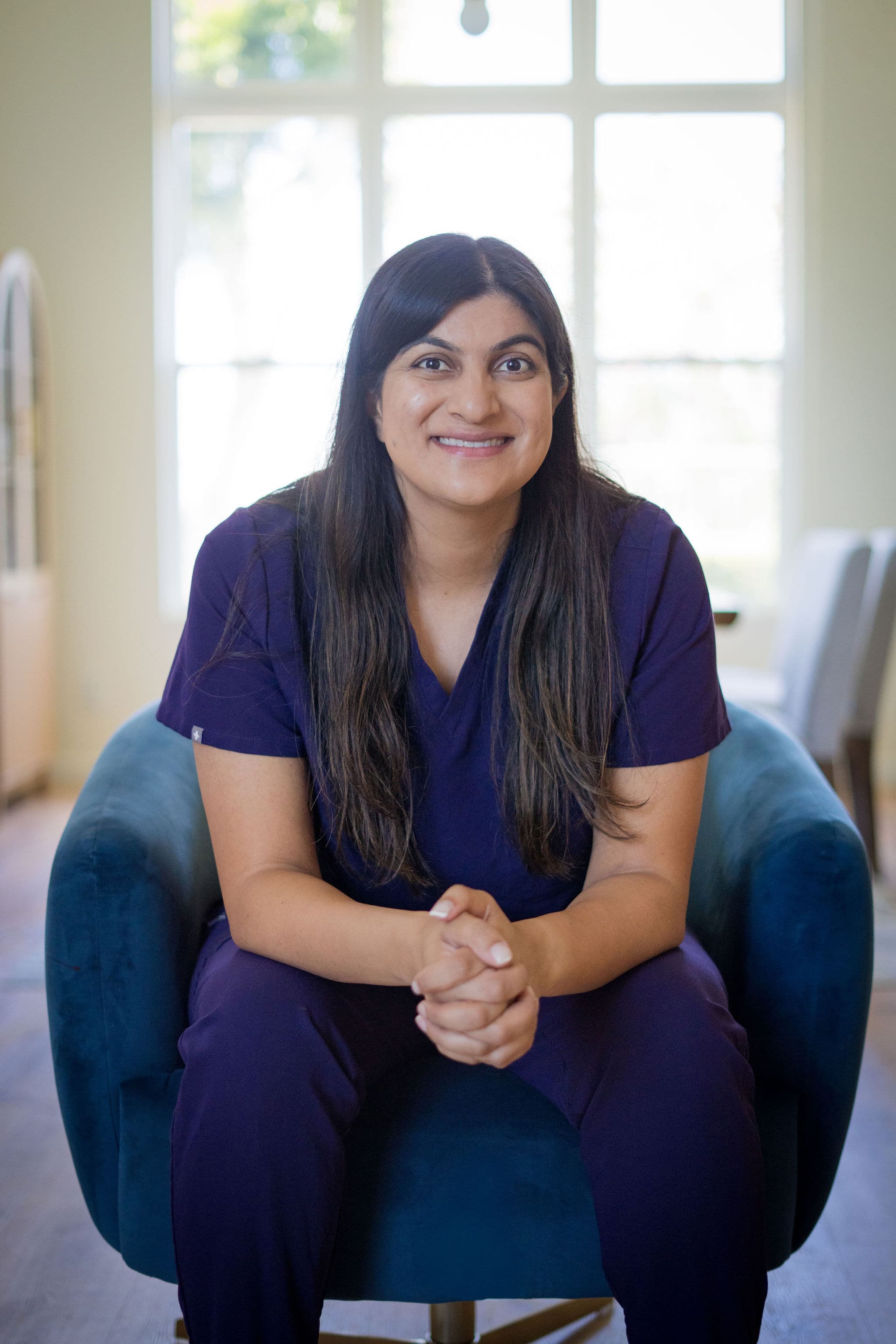 Nicki Rao, seated in an armchair by a bright window, hands clasped, smiling toward the camera.