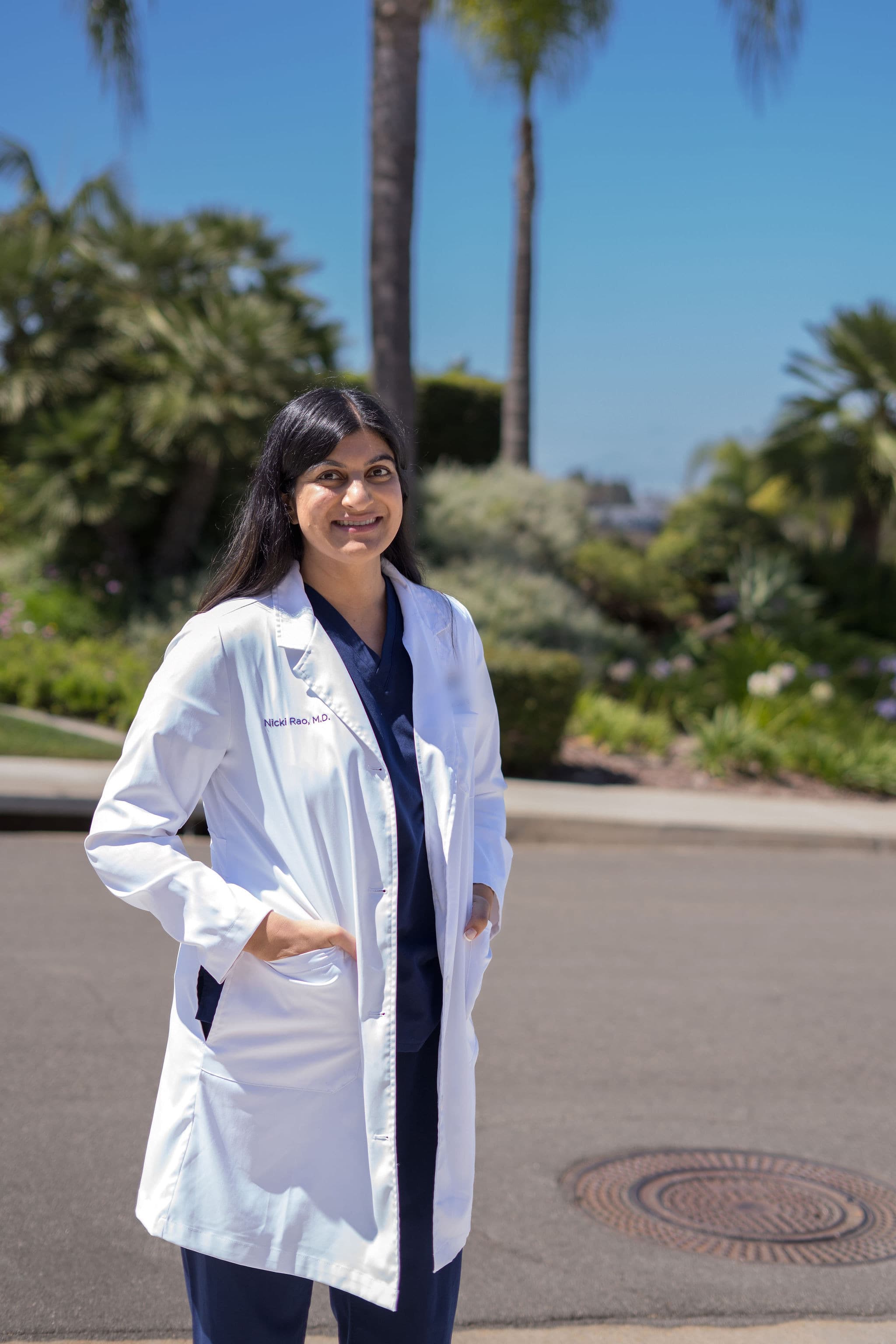 Nicki Rao outdoors in a white coat over navy scrubs, smiling toward the camera, palm trees and blue sky behind her.