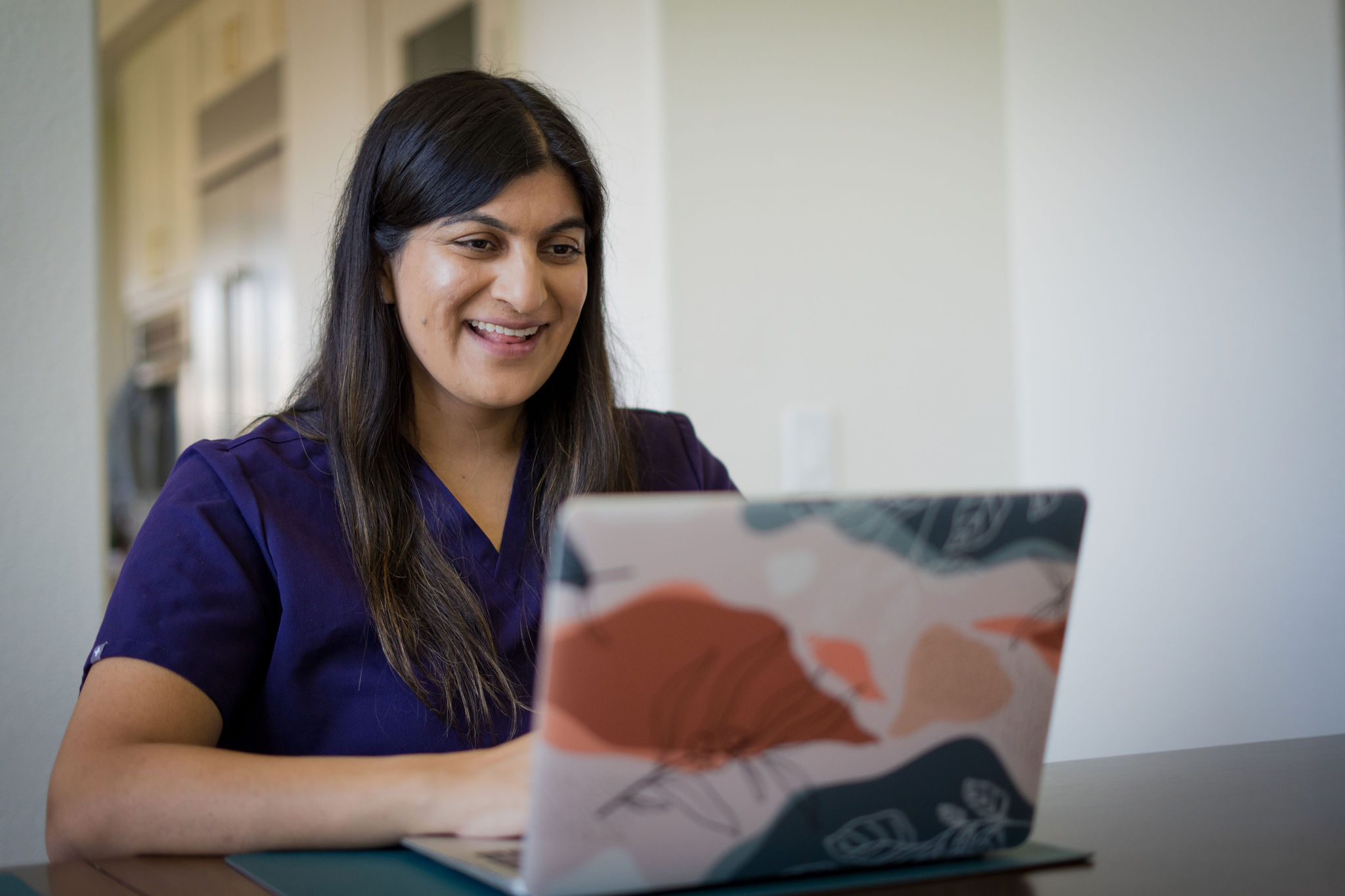 Nicki Rao at a dining table in purple scrubs, smiling at an open laptop during a video visit.